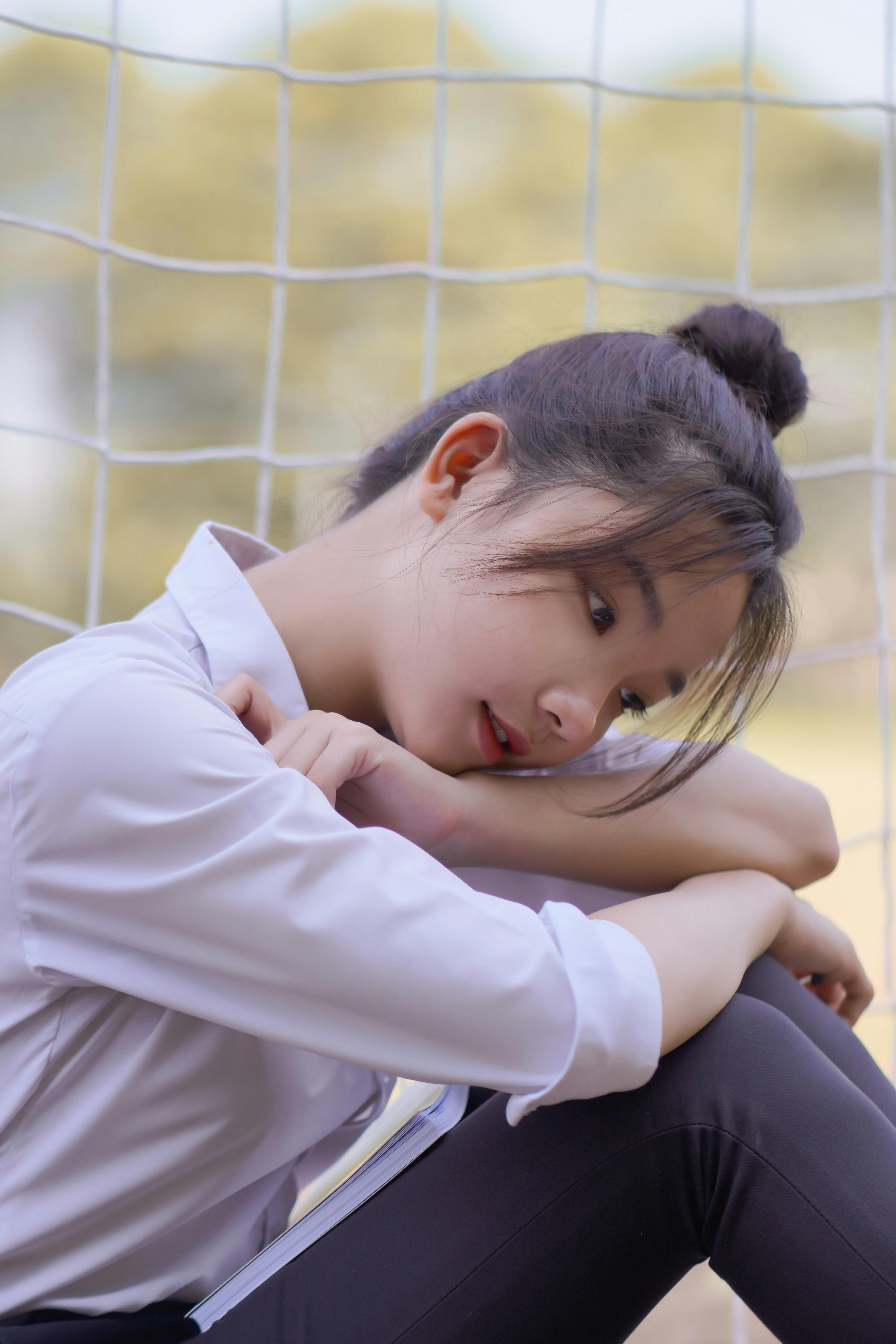 A teenage girl in uniform sits pensively outdoors by a soccer net.