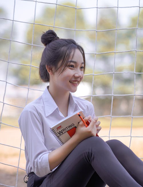 A young woman sitting on a soccer net holding a book