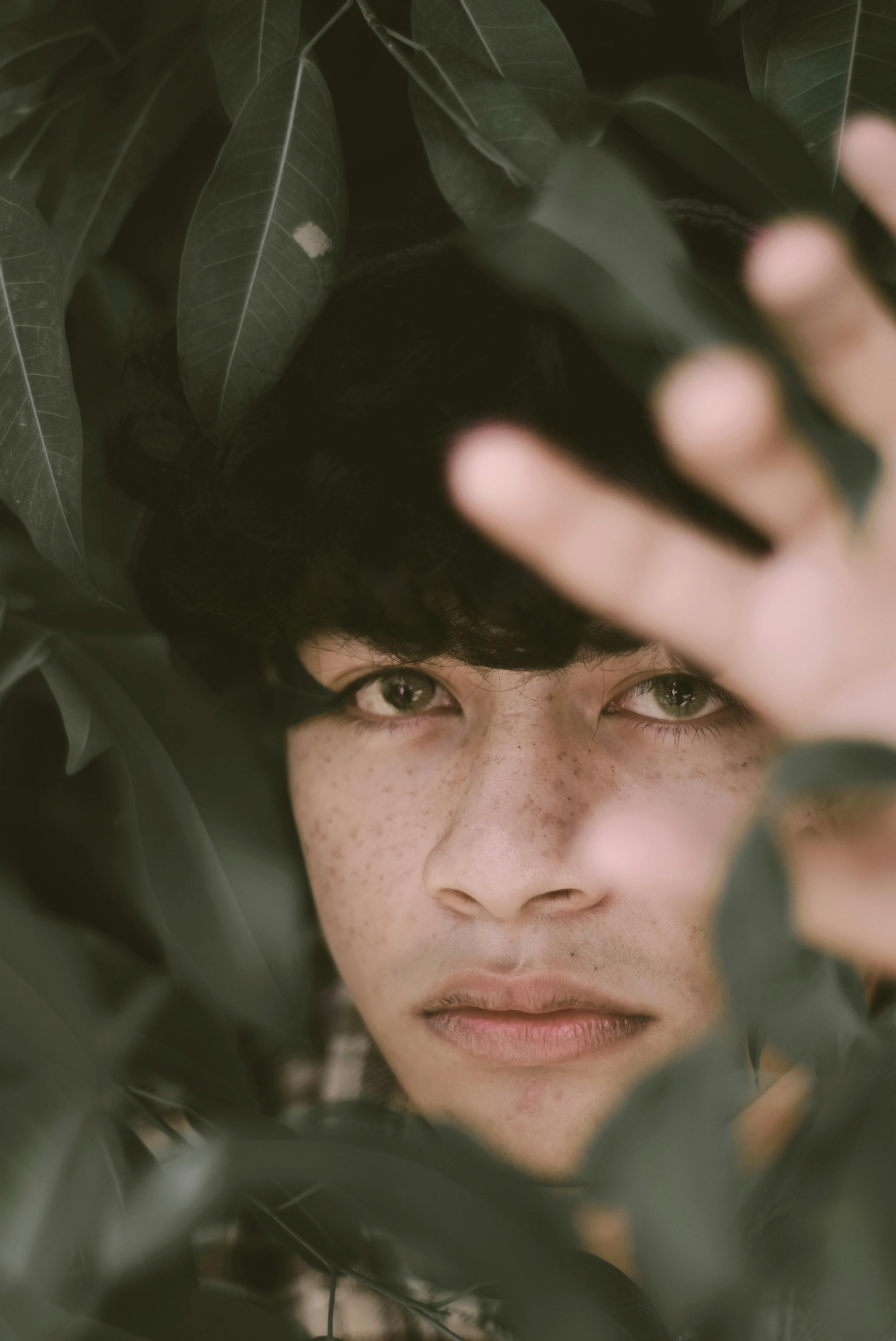 Close-up portrait of a young man surrounded by lush green leaves, creating a mysterious and natural feel.