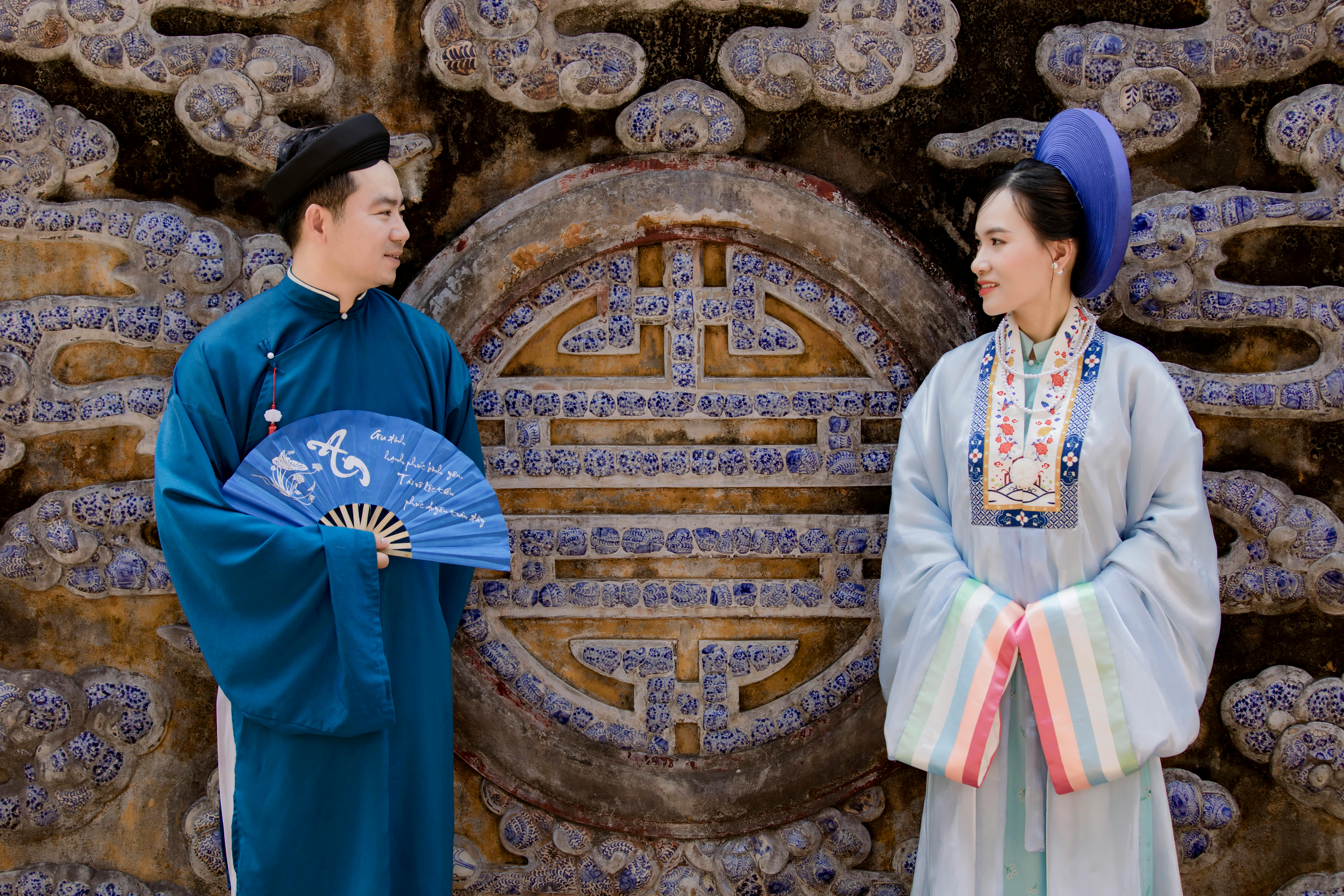Asian man and woman in traditional attire posing against a decorative wall.