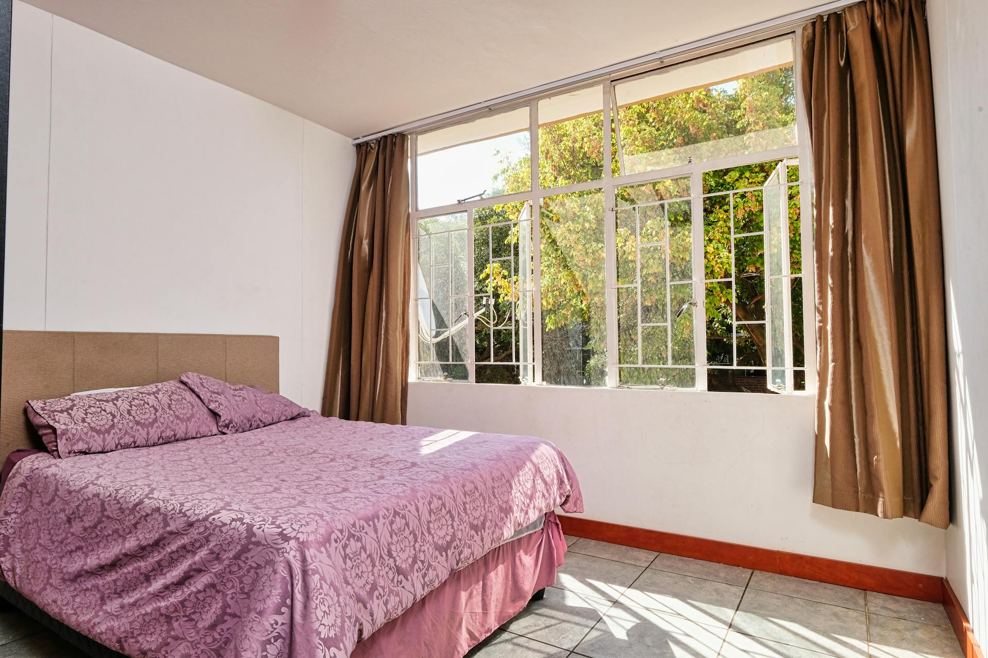 Sunlit bedroom with pink bedding and large windows overlooking a garden. Ideal for real estate listings.