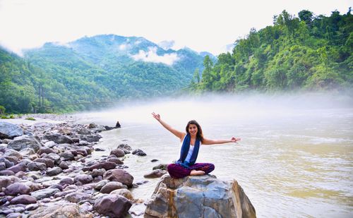 Free A smiling woman sits on a rock by a misty river, surrounded by lush mountains, enjoying nature. Stock Photo