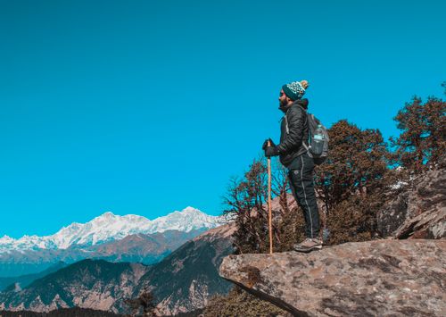 Free A hiker standing on a rocky ledge overlooking snowy mountain peaks under a clear blue sky. Stock Photo