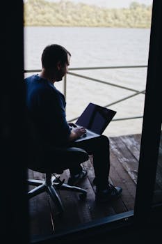 一名男子坐在湖边的甲板旁，专注而放松地使用着笔记本电脑