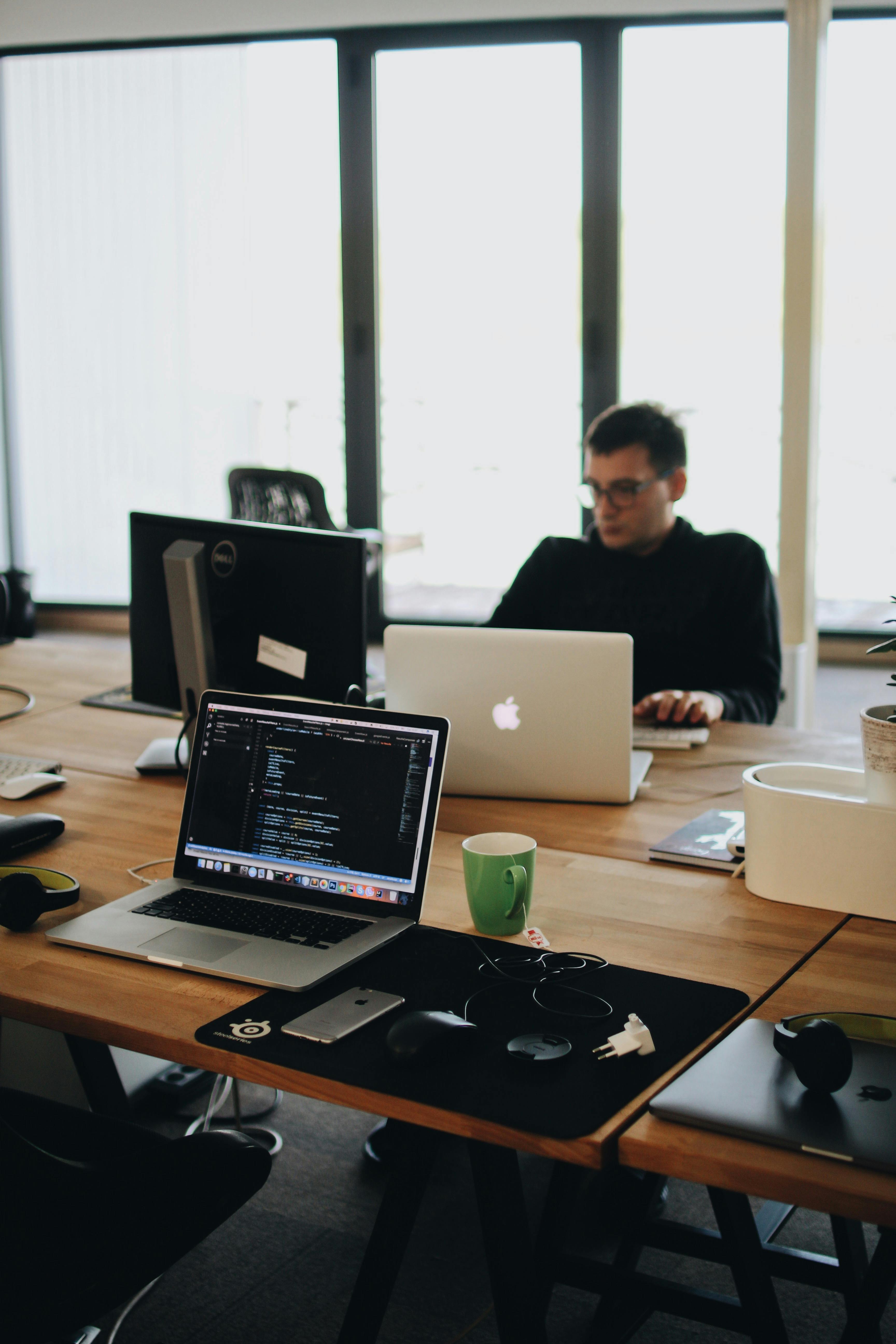A man working at a desk with two laptops and a desktop monitor in a modern office setting. One of the laptops displays lines of code. A green coffee mug is placed on the desk. Запитати в ChatGPT