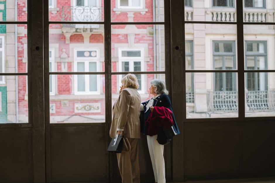 Dos mujeres mayores observan edificios arquitectónicos a través de grandes ventanas interiores.