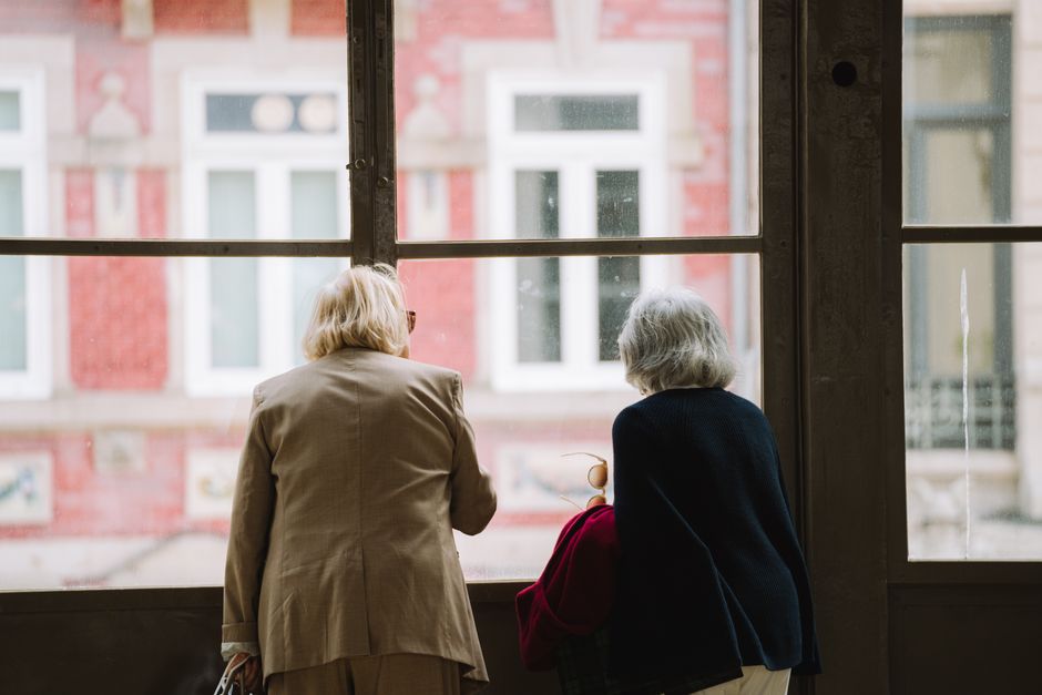 Dos mujeres mayores con cabello gris están mirando por la ventana a una escena urbana, capturando un momento sereno.