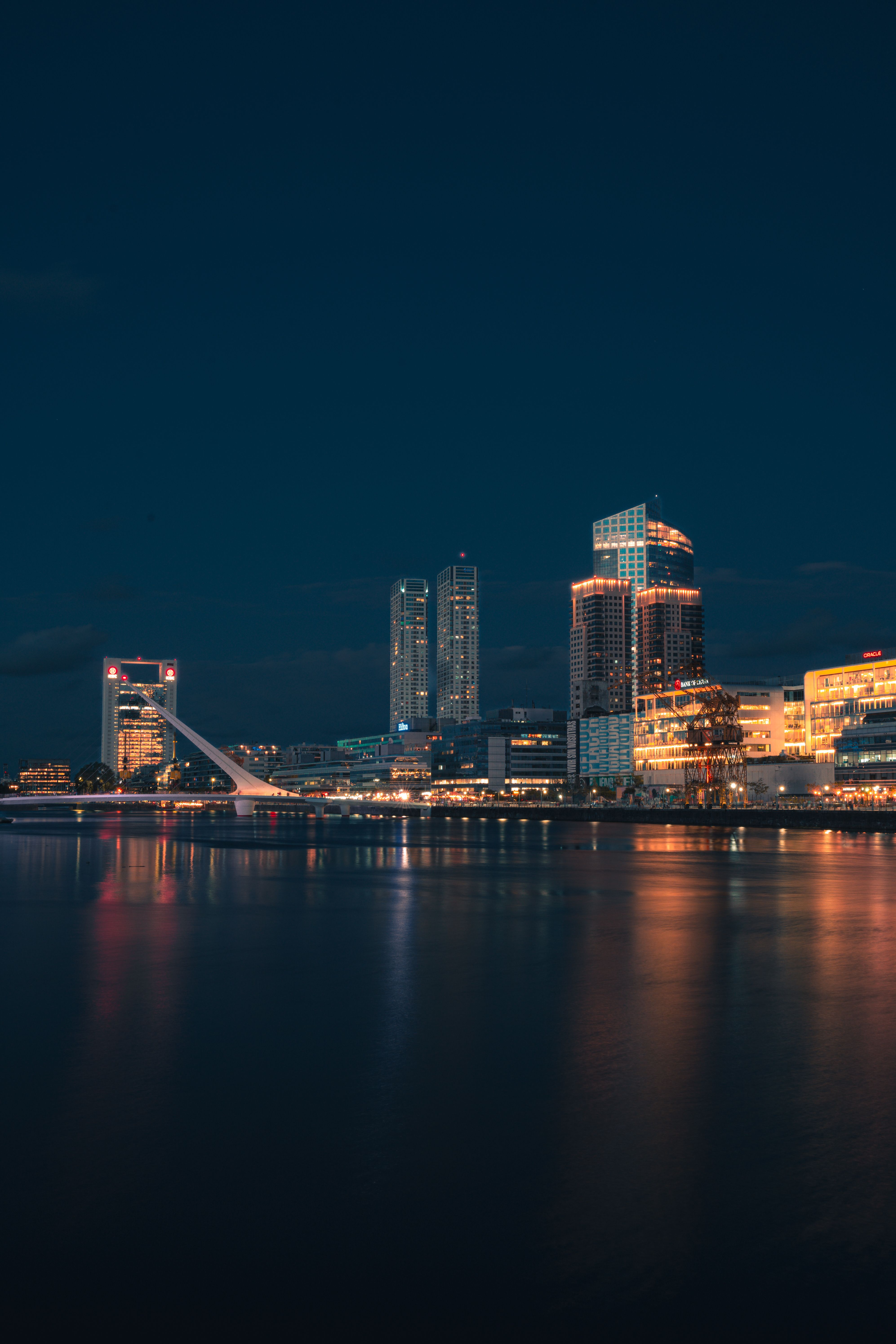 City skyline reflecting on a calm river at night, with illuminated skyscrapers and an iconic bridge.