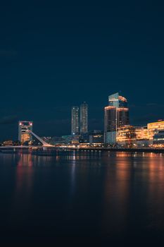 City skyline reflecting on a calm river at night, with illuminated skyscrapers and an iconic bridge.
