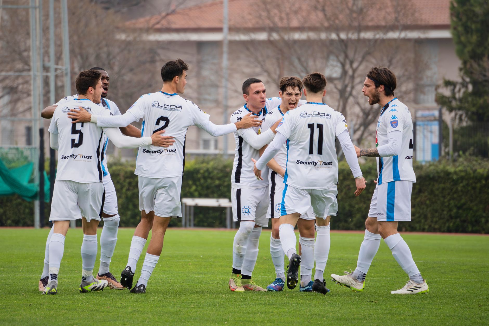 A group of soccer players in white uniforms celebrating on a soccer field during a daytime match.