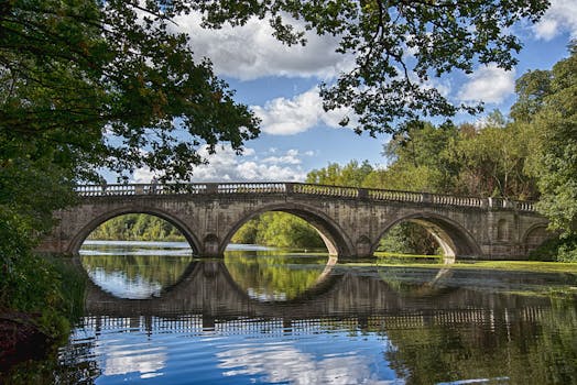 Ancient stone bridge arches over a calm river, surrounded by lush greenery and clear skies.