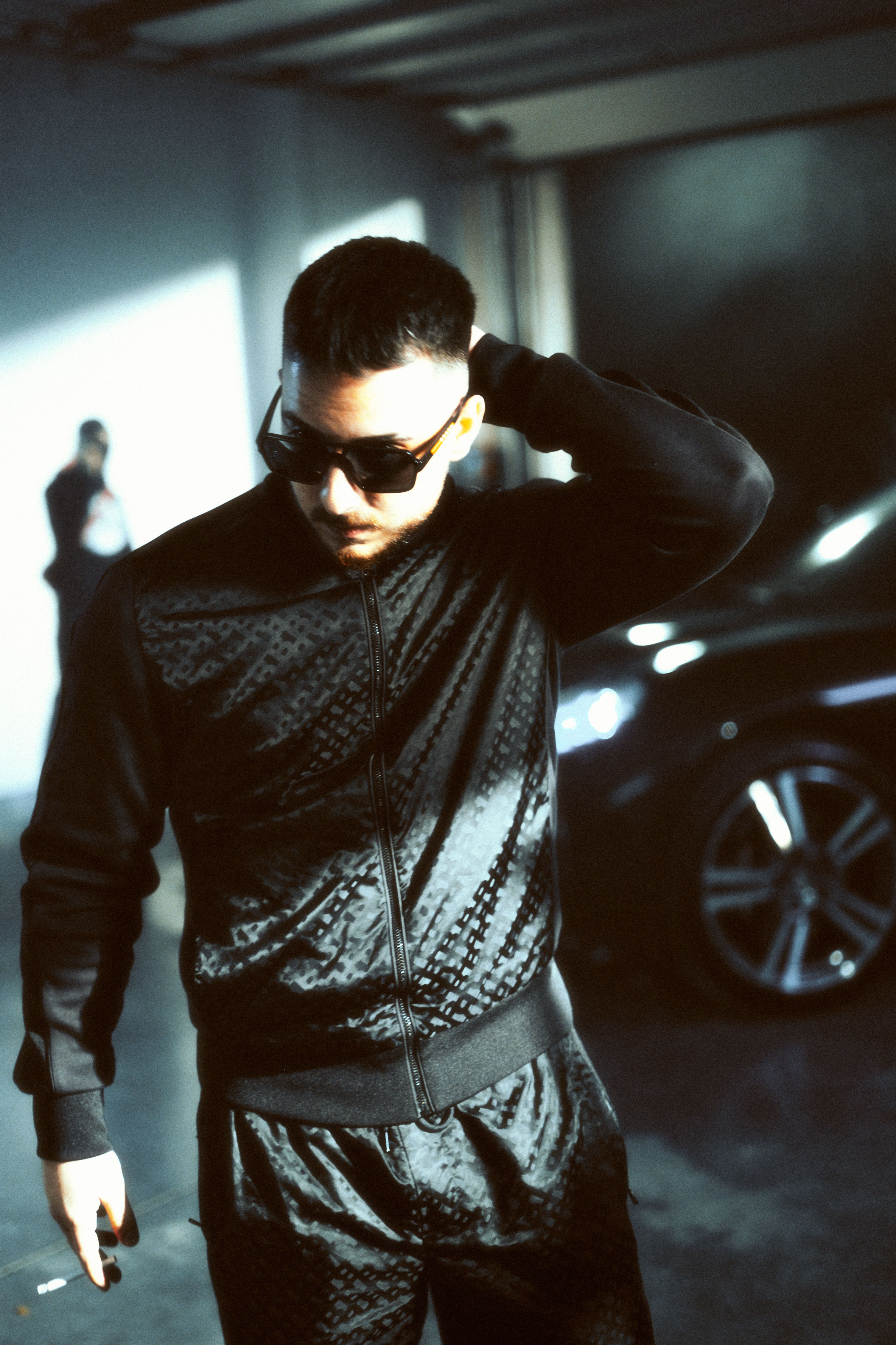 A cool and confident man posing in an urban setting with sunglasses and a black jacket.