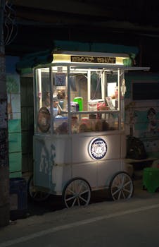 A brightly lit street food cart with wheels stands at night on an urban street.