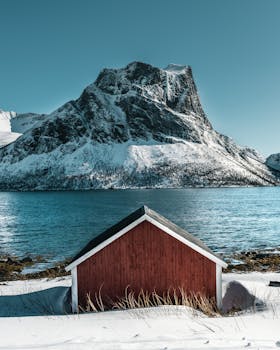 峡湾旁，一座红色小屋坐落在白雪皑皑的群山环抱中，构成一幅宁静的冬日景象