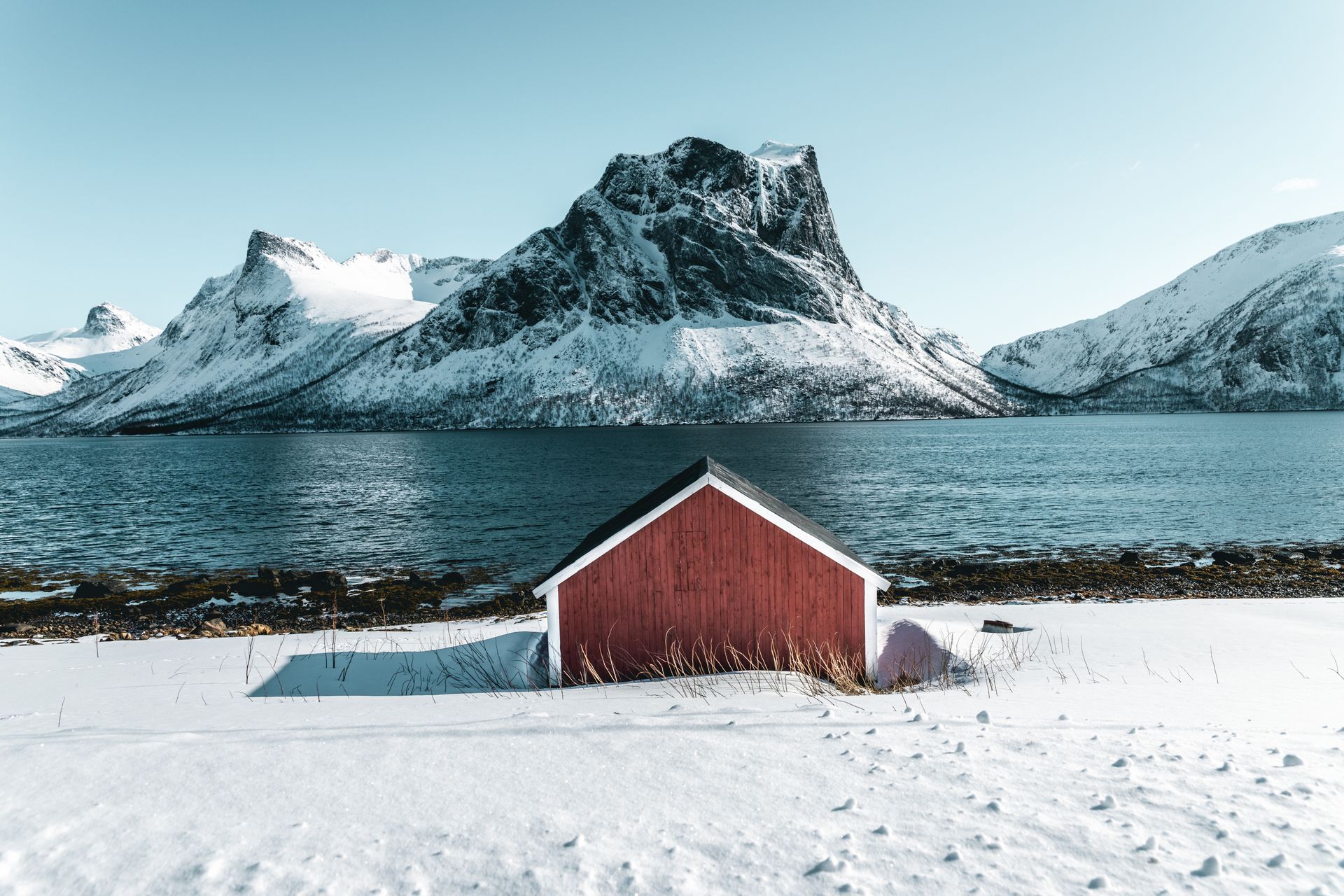 Snow-covered mountains and lake with a red barn create a tranquil winter landscape scene.