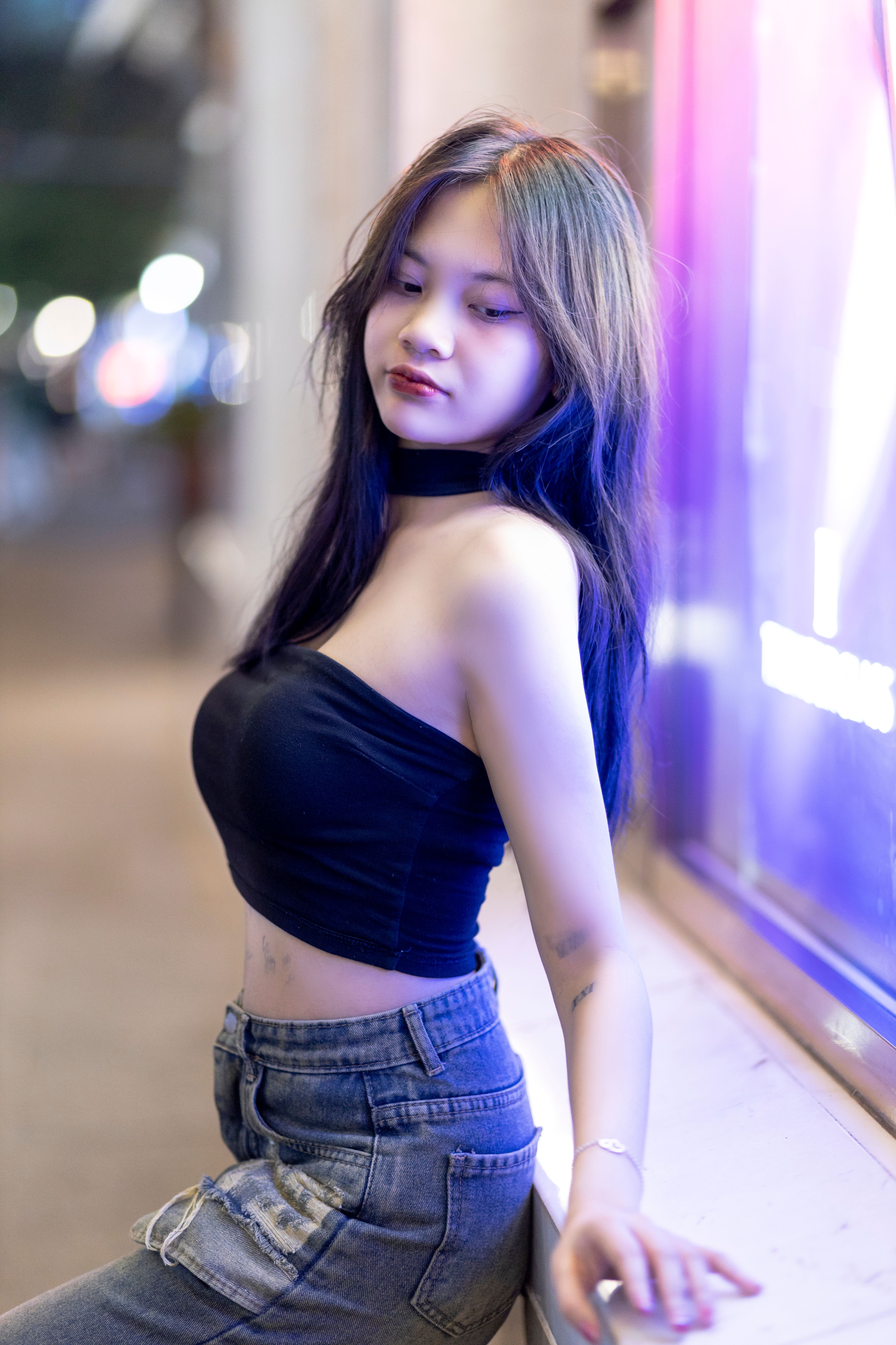 Fashionable woman in a black bandeau top and jeans posing against a vibrant urban backdrop at night.