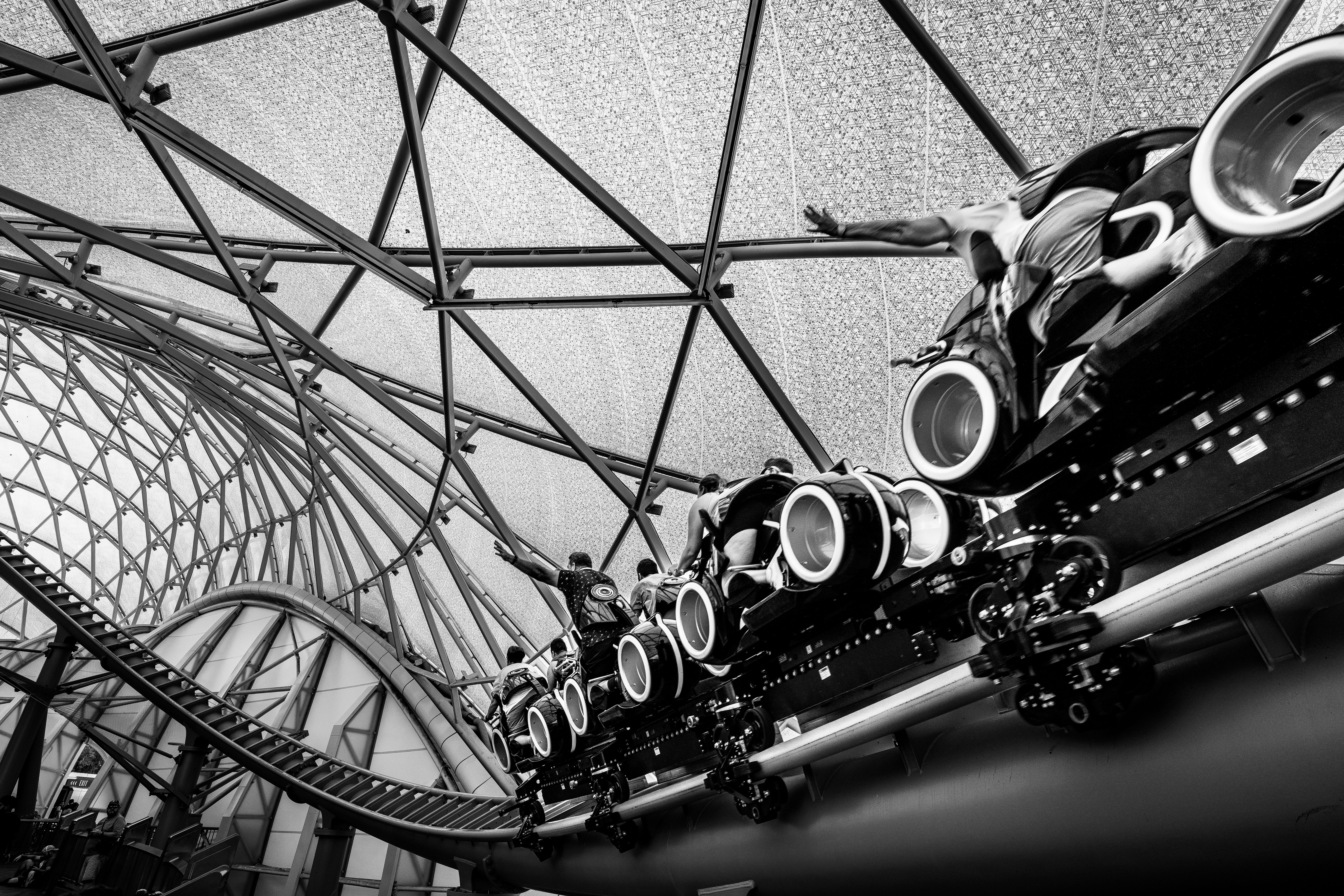 Black and white image of a roller coaster ride under a glass roof structure, capturing the excitemen
