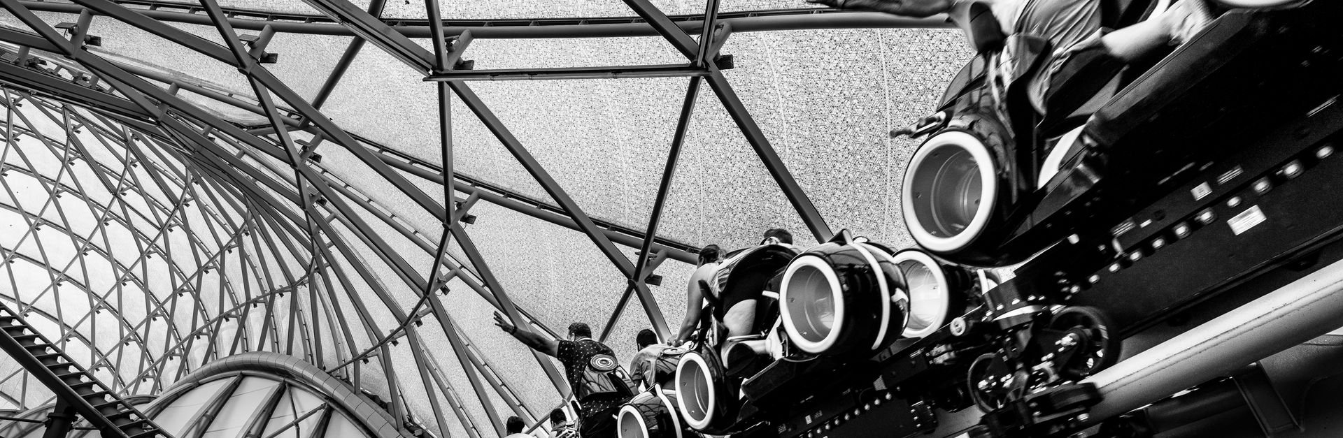 Black and white image of a roller coaster ride under a glass roof structure, capturing the excitemen