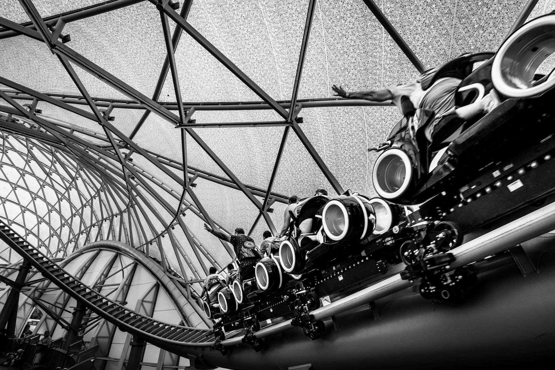 Black and white image of a roller coaster ride under a glass roof structure, capturing the excitemen