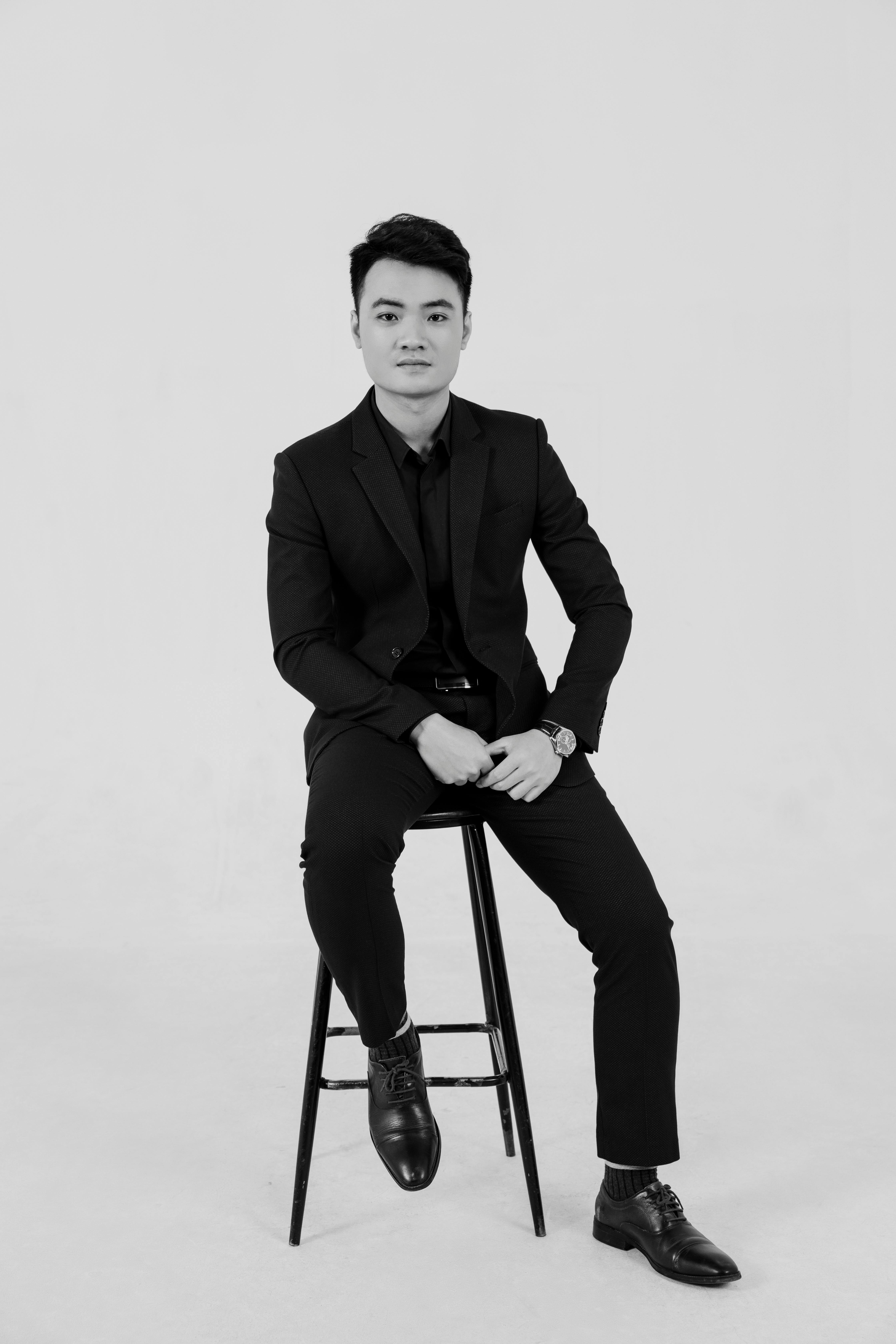 Portrait of a man in an elegant black suit seated on a stool in a studio setting.