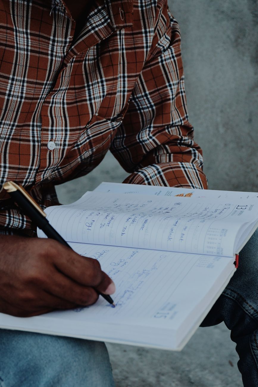 Une personne portant une chemise à carreaux marron écrit avec un stylo noir dans un agenda ouvert.