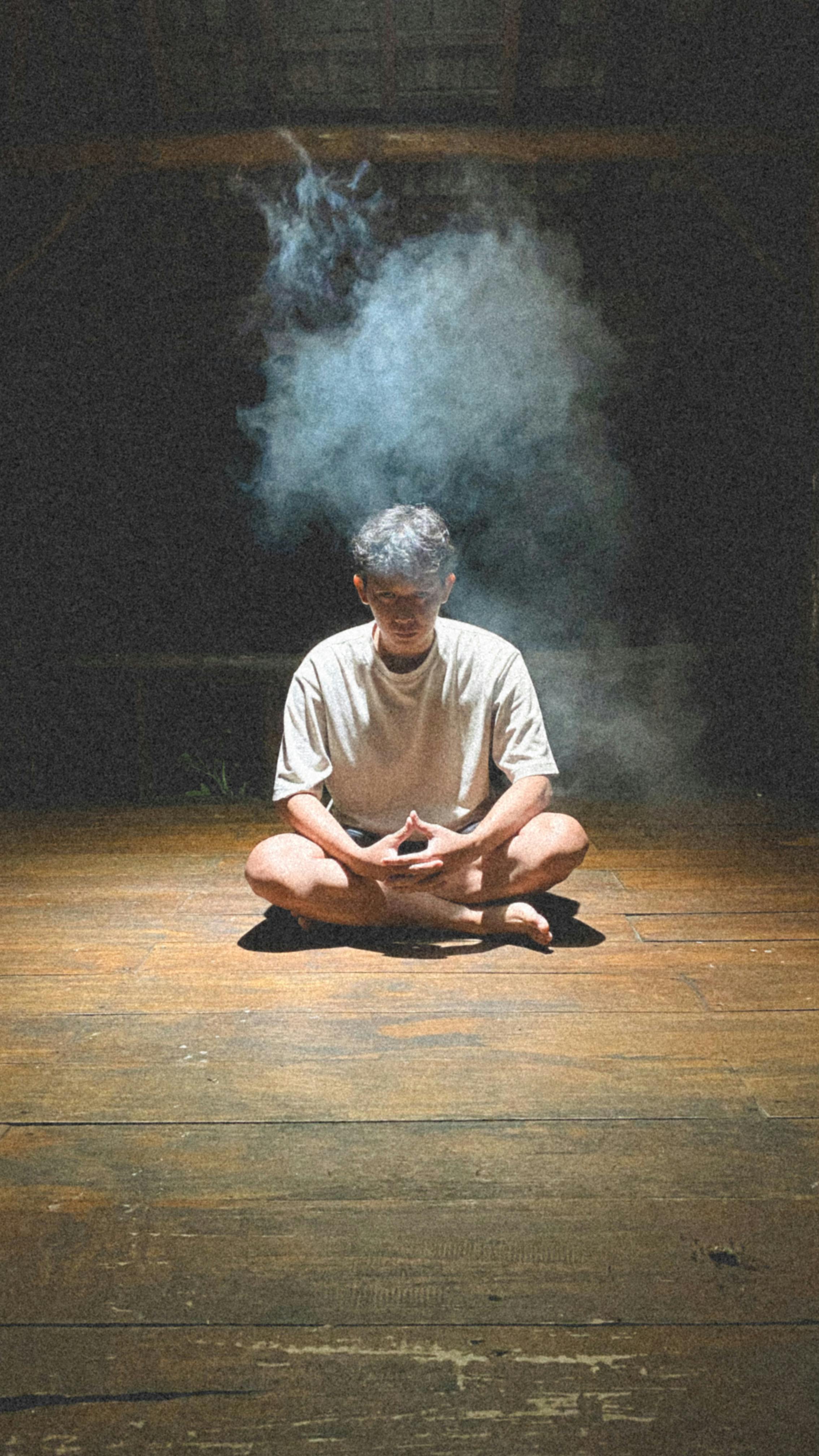 A man meditating alone in a dimly lit attic with smoke surrounding him.