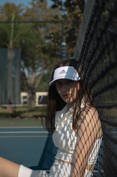 Woman sitting against tennis net, in fashionable sports attire and visor.