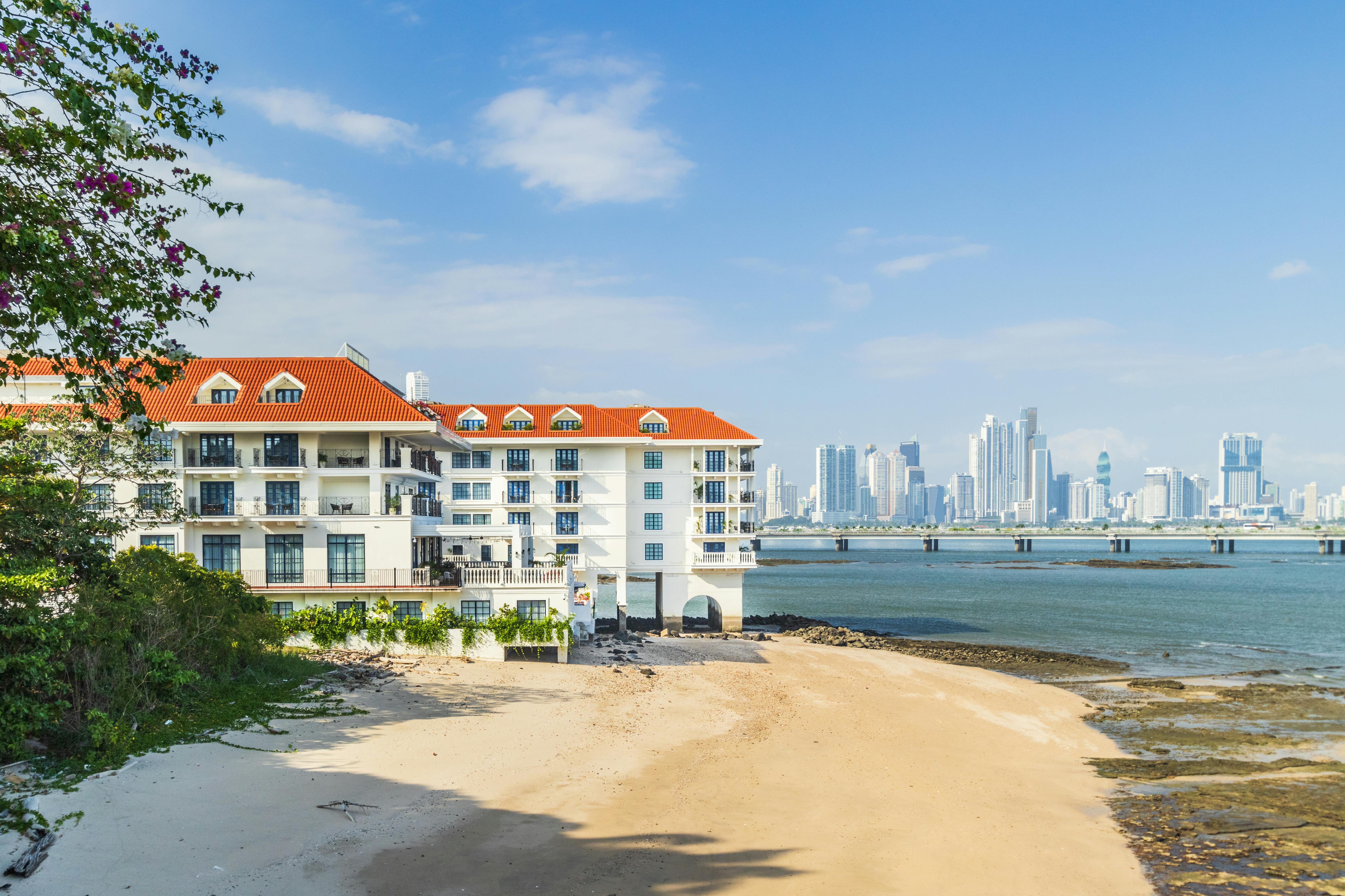 White beachfront hotel overlooking Panama City skyline and ocean under a blue sky.