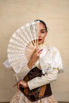 Young woman in traditional dress with lace fan in elegant pose against beige backdrop.