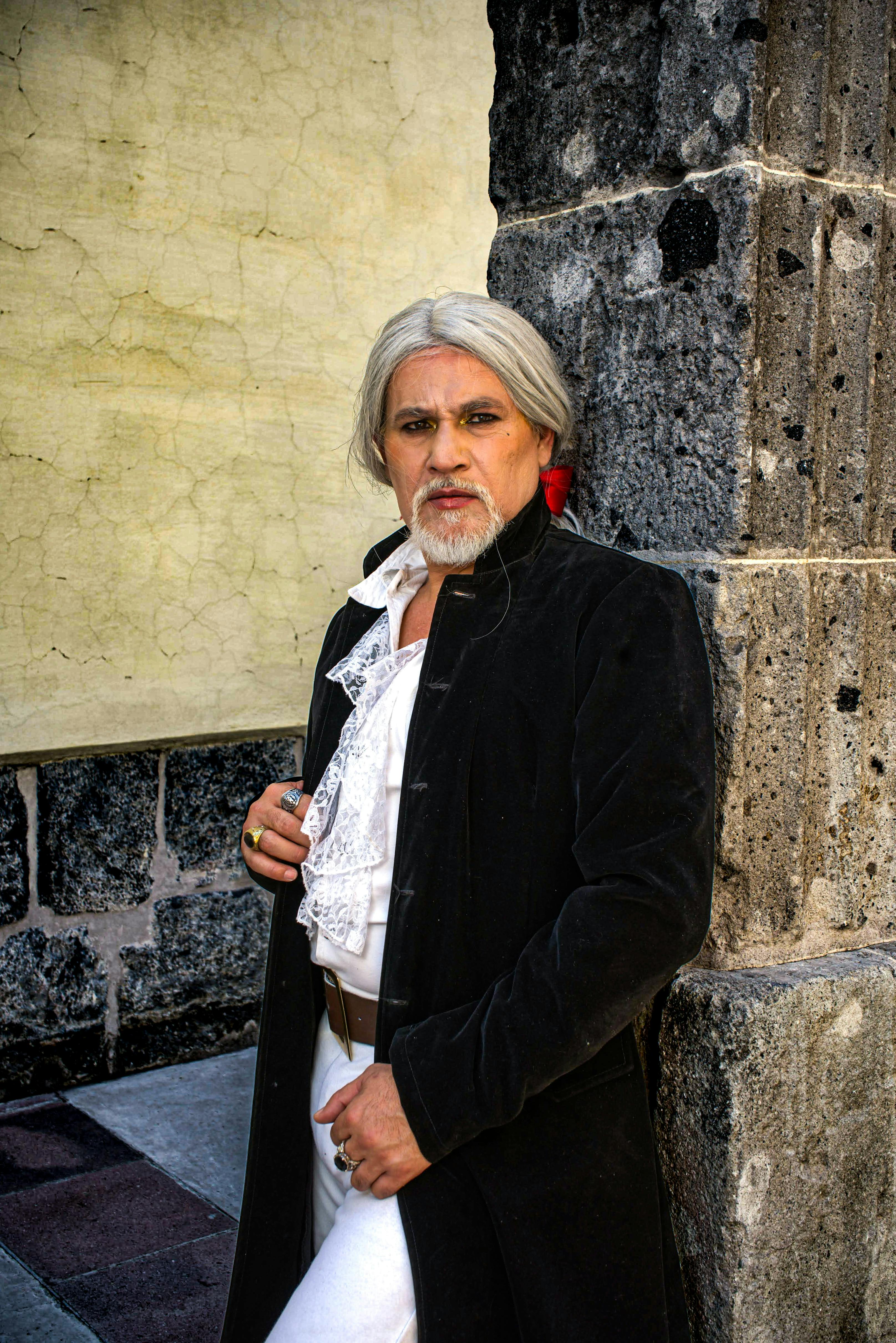 Elderly man wearing a vintage costume poses against a stone wall, evoking a historical vibe.
