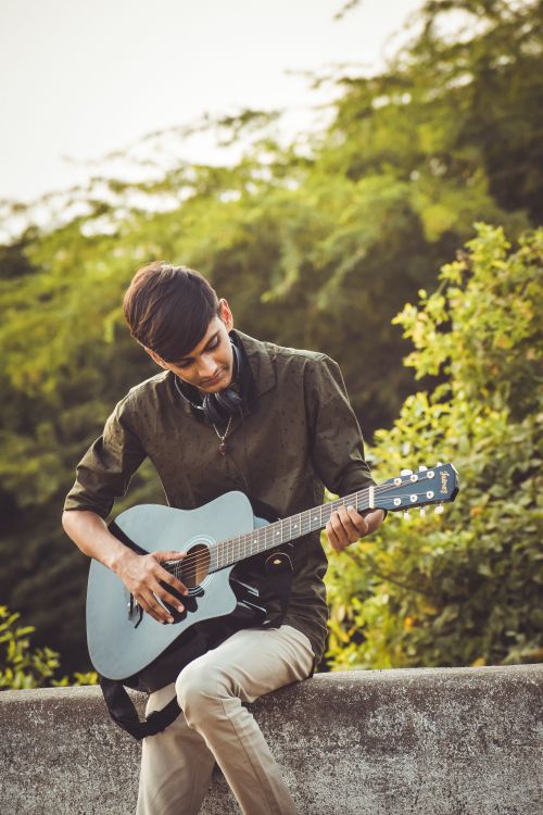 Free A young man plays guitar while sitting outdoors in a sunny park, enjoying music and leisure time. Stock Photo