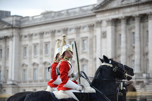 Two people in red uniforms riding horses in front of buckingham palace