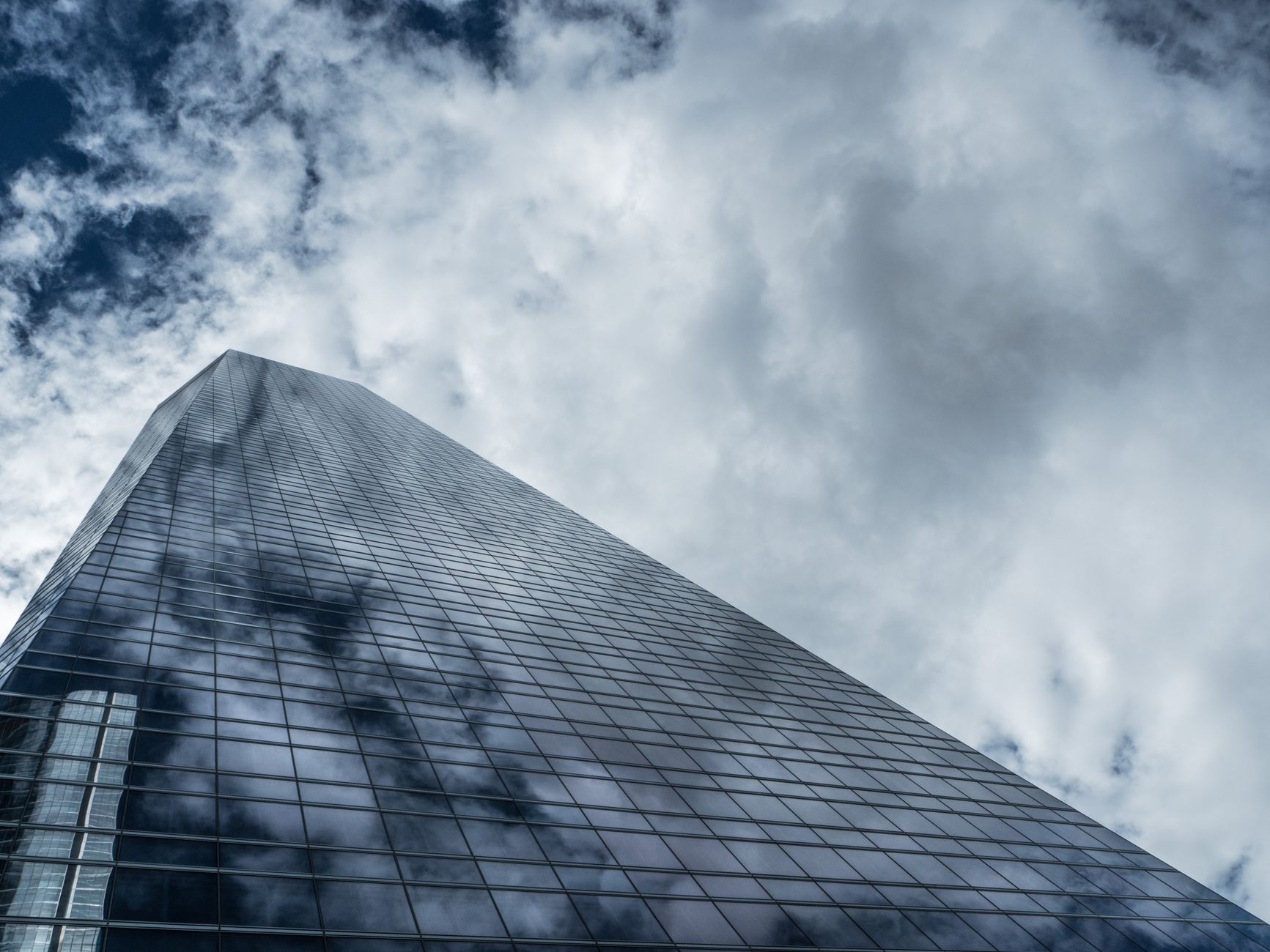 Low angle view of a towering modern skyscraper with glass windows reflecting clouds against a dramatic sky.