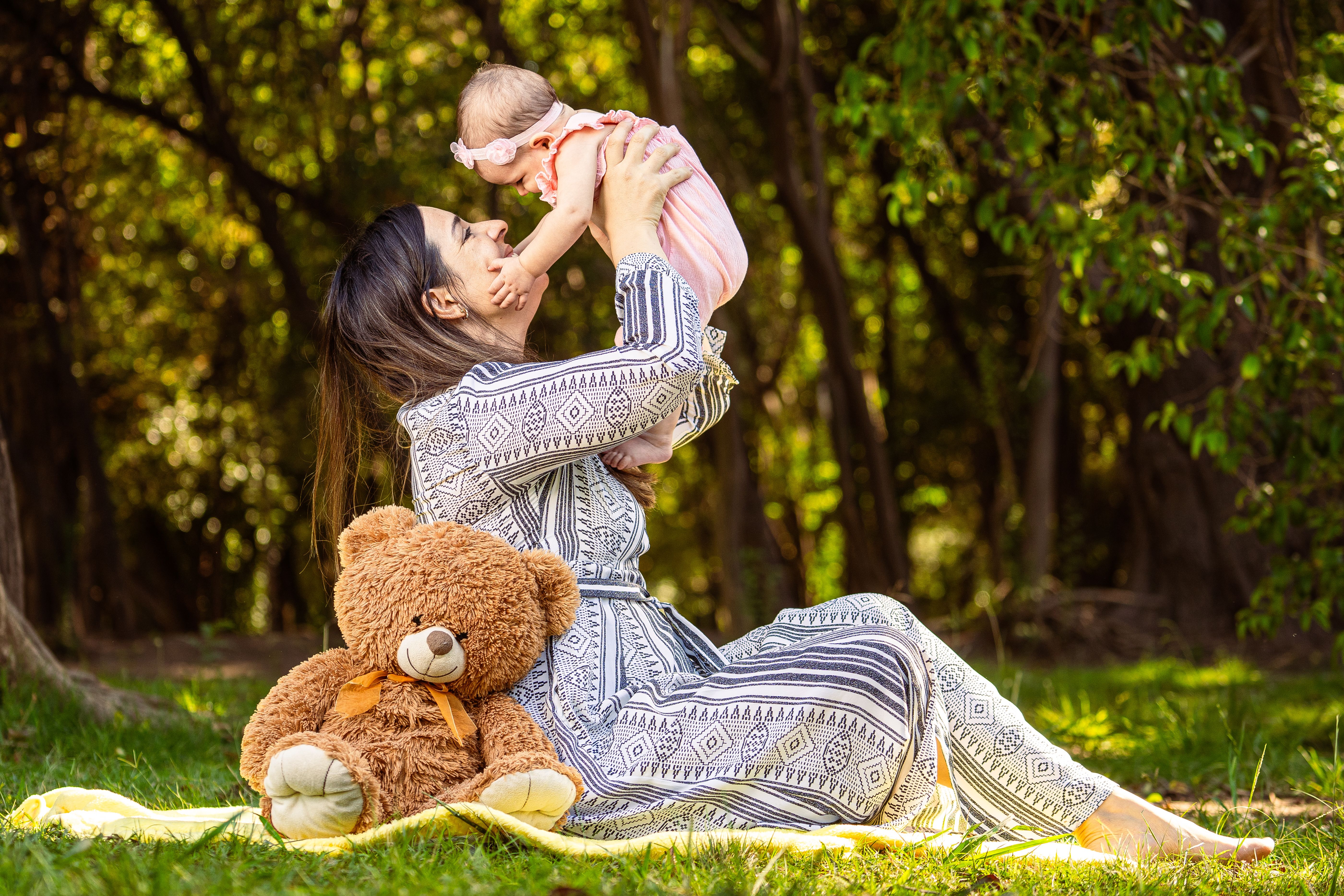 A joyful mother lifting her baby in an outdoor setting with a teddy bear nearby.