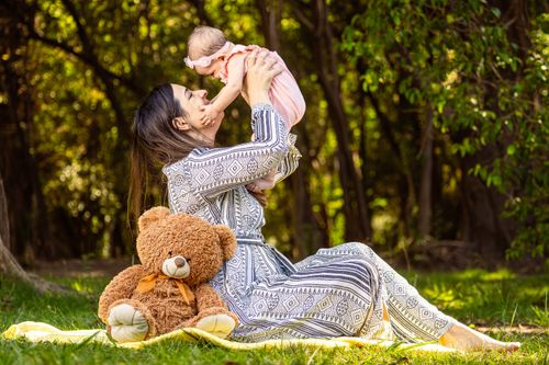 Free A joyful mother lifting her baby in an outdoor setting with a teddy bear nearby. Stock Photo