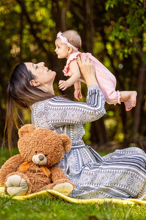 Free A joyful mother lifts her baby outdoors, surrounded by nature and a teddy bear. Stock Photo