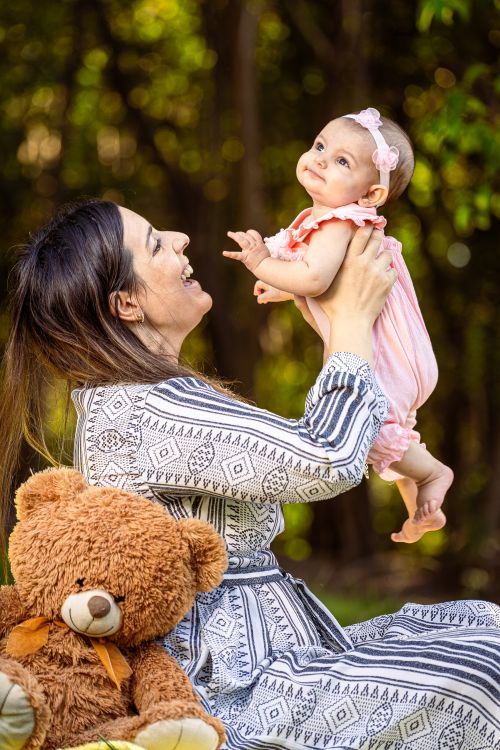 Free A mother joyfully lifts her smiling baby girl outdoors, with a teddy bear by their side. Stock Photo