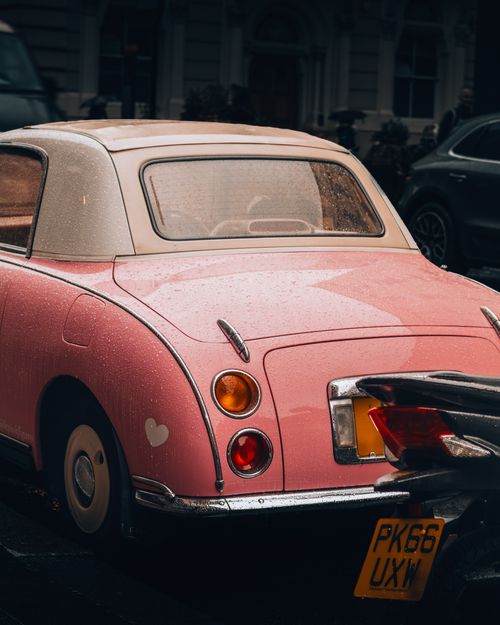 Free Charming vintage pink Nissan Figaro car parked in a rainy urban street, showcasing retro aesthetics. Stock Photo