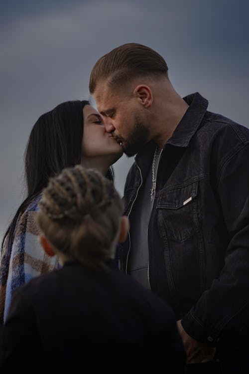 A man and woman kiss their child while standing in front of a cloudy sky