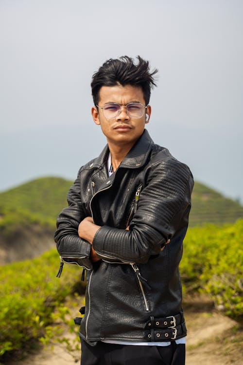 A young man in glasses and leather jacket standing in front of a green hill