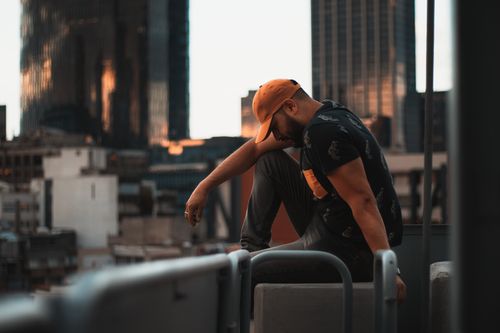 Free A man wearing a cap, sitting pensively on a rooftop with a city skyline at sunset. Stock Photo