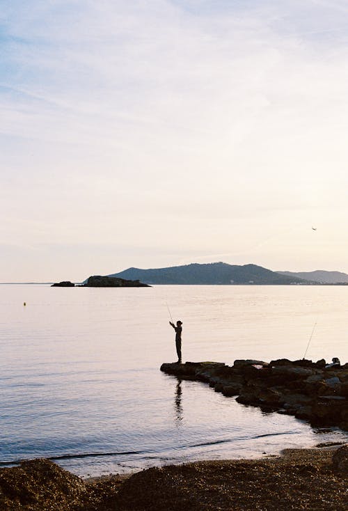 Free Silhouette of a Person Fishing on a Shore in the Evening  Stock Photo