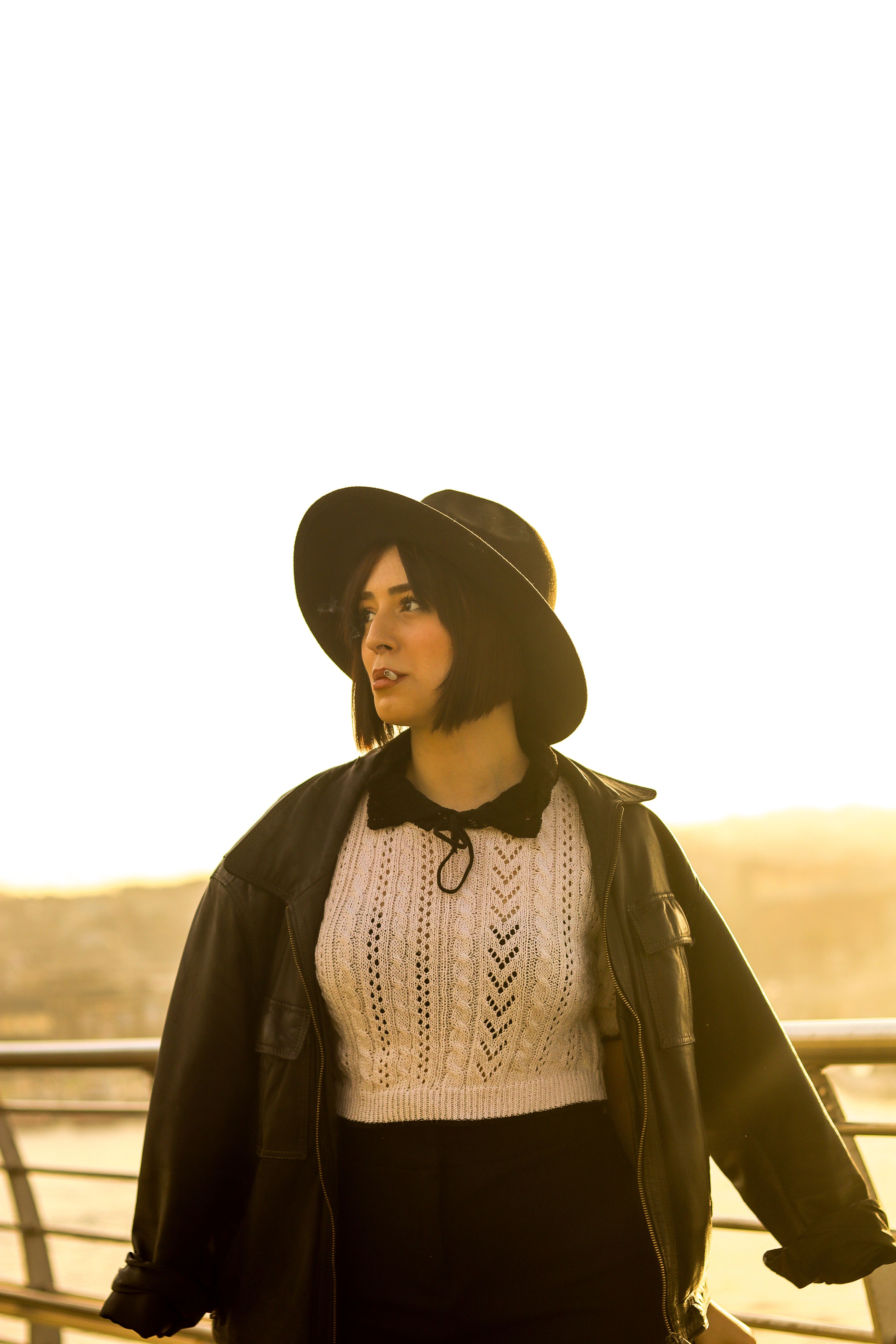 Young woman in a hat and jacket poses stylishly against a sunset backdrop.
