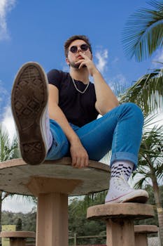 一个戴着墨镜、穿着牛仔裤的年轻人坐在户外的桌子旁，散发着轻松的气质