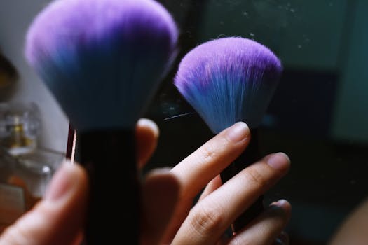 Close-up of a hand holding a purple-bristled makeup brush, reflecting in a mirror.