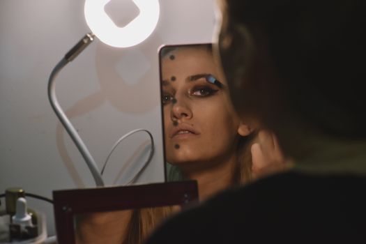 A woman applies makeup using a mirror under soft lighting, captured in a reflective portrait setting.