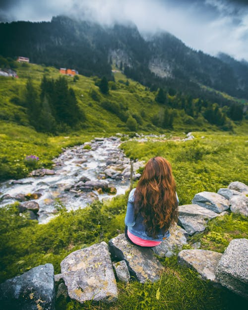 Woman in Denim Jacket Sitting on Rock Facing Mountain