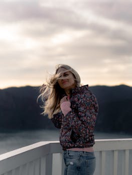 一位身穿花卉夹克的金发女子站在风景优美的阳台上，背景是连绵的山脉，感受着微风拂面