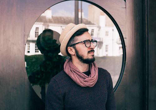 Free Portrait of a fashionable man wearing a hat and scarf, standing outdoors with a reflective background. Stock Photo