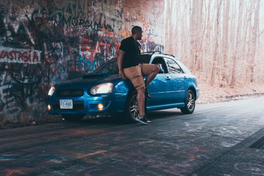 Man posing with a blue car near a graffiti-covered tunnel in a wooded area, Williamsburg, VA.